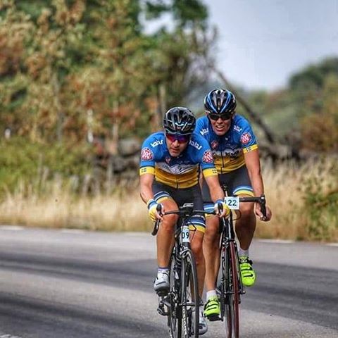 Foto på två personer som cyklar på en landsväg. De har på sig cykelhjälm och blå-gula träningskläder.