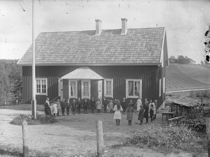 Ett äldre foto på en skolklass utanför ett äldre skolhus. Barnen står uppställda i nästan en hel ring framför huset.