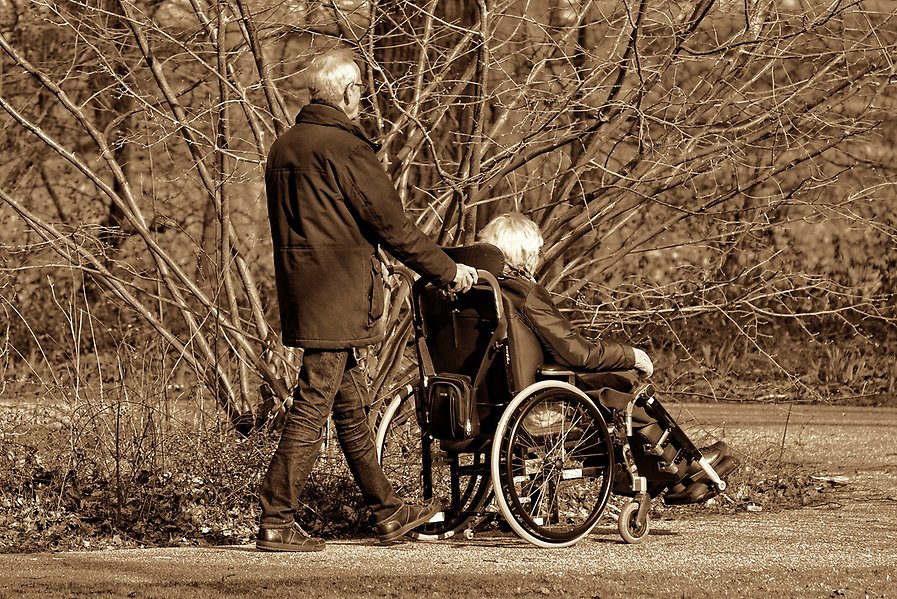 En äldre man kör en rullstol i vilken en äldre kvinna sitter.