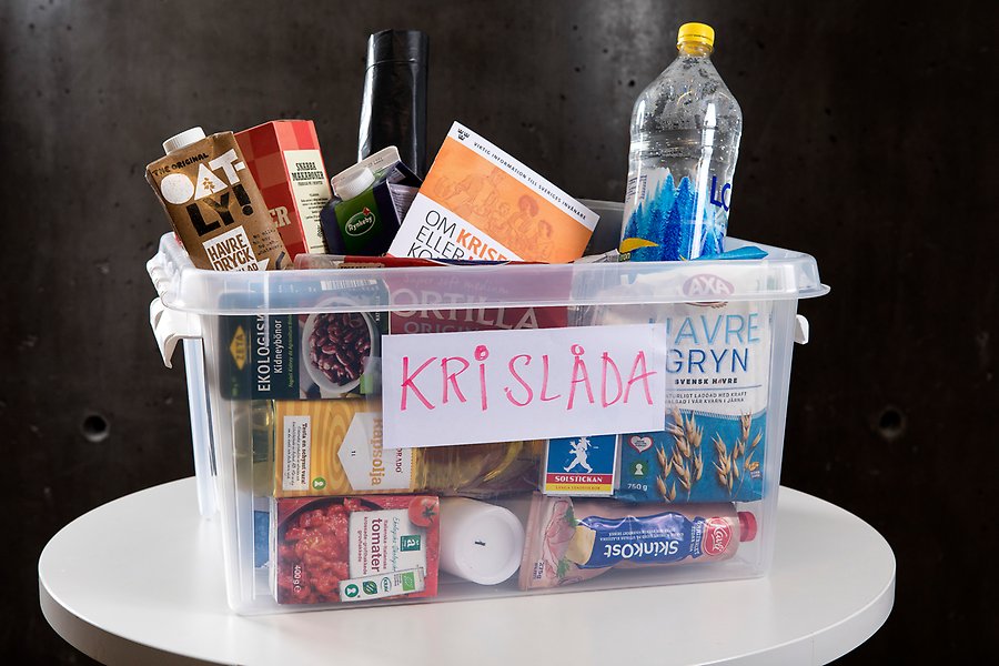 Foto på en låda fylld med mat, vatten och broschyren om Kriget eller krisen kommer. På lådan står det Krislåda i versaler.