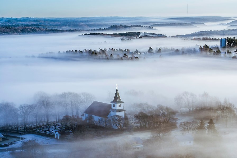 Flygfoto över Foss kyrka under vintertid. Dimman ligger som en slöja över omliggande mark med skog och den vita kyrkan tornar upp sig över omgivningen.