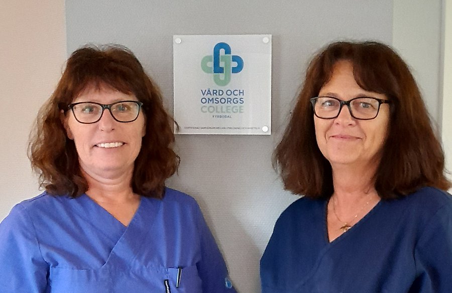 Två kvinnor i blå kläder som ler mot kameran. På väggen bakom dem syns en skylt där det står vård- och omsorgscollege.