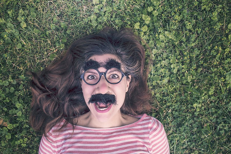 Ung kvinna med lösnäsa med mustasch och glasögon med lösögonbryn. Hon ligger på en gräsmatta.