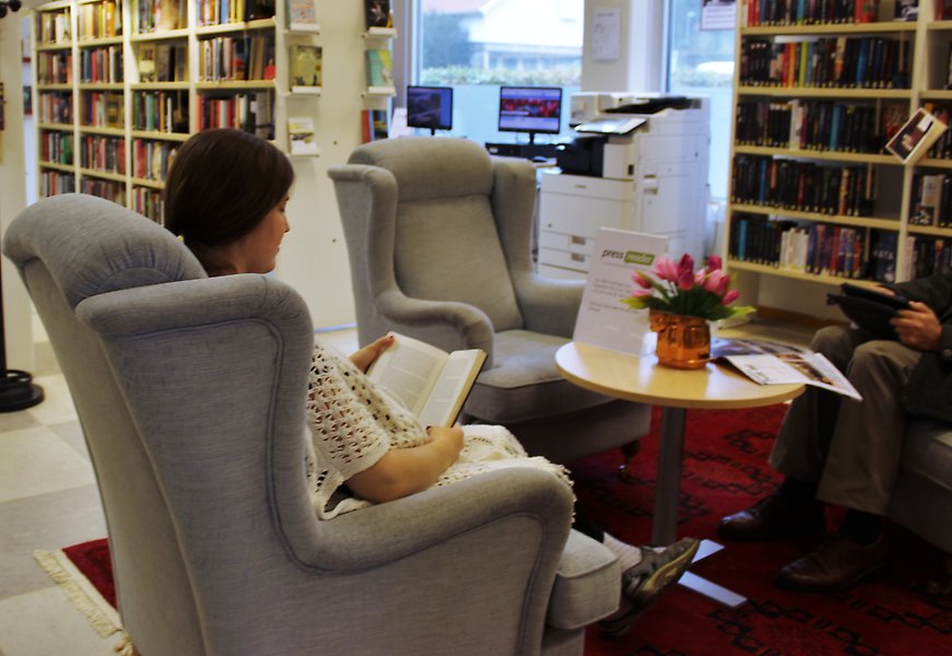 Foto på två personer som sitter i fåtöljer i ett ljust bibliotek. Den ena personen läser en bok och den andra läser på en surfplatta.