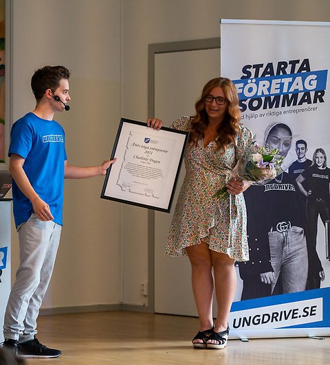 En person som ger ett stort inramat diplom till en annan person. I bakgrunden syns en roll-up där det står Starta företag i sommar och ungdrive.se.