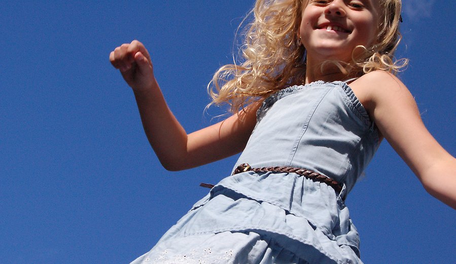 Bild på en blond flicka med lockigt hår som hoppar upp i luften, i bakgrunden syns en klarblå himmel.
