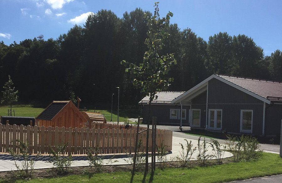 Solig bild på utsidan av Önnebacka förskola som är en mörkgrå träbyggnad med rött tegeltak.