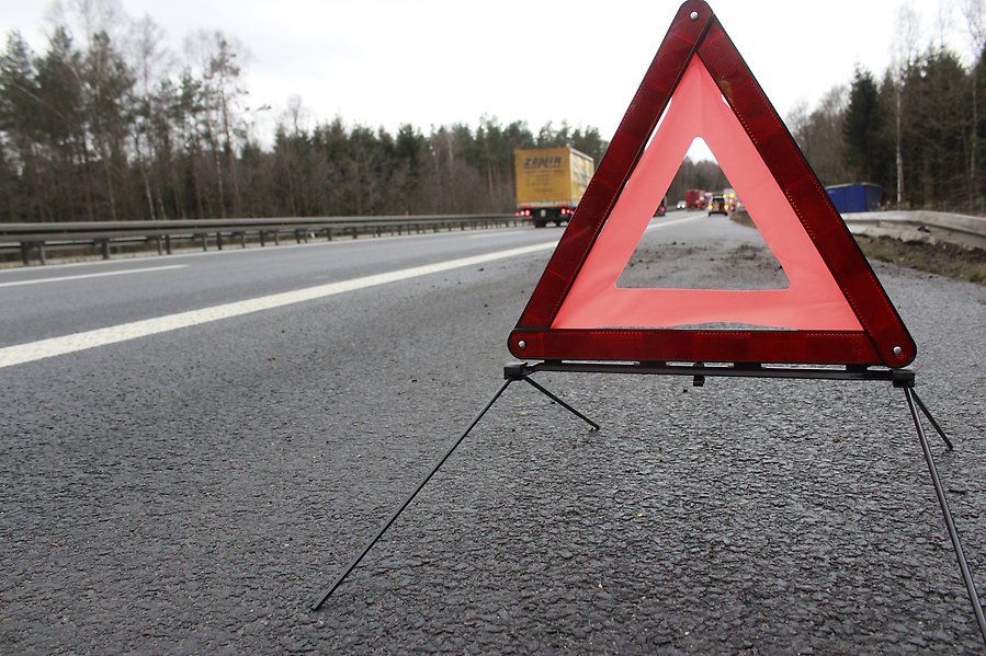 Närbild av en varningstriangel som står på en asfaltsväg. I bakgrunden syns räddningsfordon.