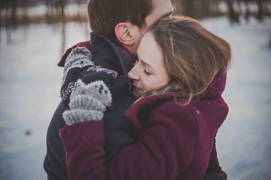 En man och en kvinna kramar varandra. Kvinnan har en vinröd kappa på sig och de står utomhus en solig vinterdag.