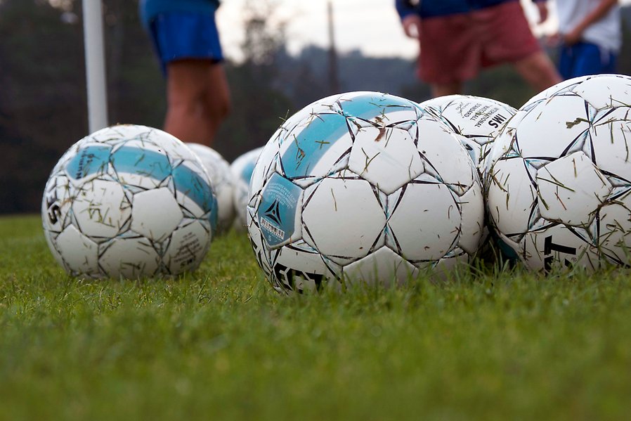 Foto på flera vita fotbollar på en grön gräsmatta. I bakgrunden syns ben på fotbollsspelare.