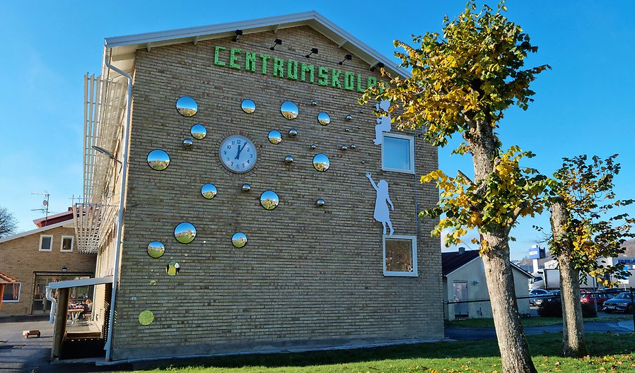Foto på Centrumskolan i Dingle. En byggnad i gult tegel med texten "Centrumskolan" i grönt och ett konstverk med silvriga bubblor. I förgrunden en grön gräsmatta och träd med höstlöv. Det är solsken och klarblå himmel.