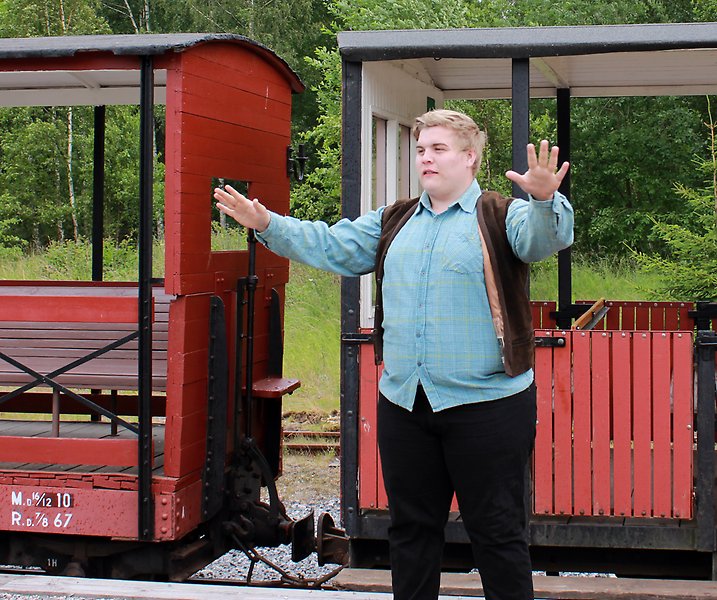 Foto på en person som står på en perrong vid ett mindre öppet tågset. Personen har på sig en jeansskjorta och en väst och håller upp armarna och gestikulerar. 
