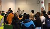 I en lektionssal står cirka femton elever och tittar på en lärare som pekar på en whiteboardtavla.