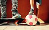 Närbild på två barns ben och fötter. De har träningsskor på sig, den ena har en fot på en skateboard och den andre har en fot på en fotboll.