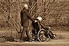 En äldre man kör en rullstol i vilken en äldre kvinna sitter.