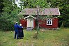 Foto på ett par i gammaldags blåa kläder som står med ryggarna mot kameran och tittar mot ett rött litet torp som ligger i skogen.