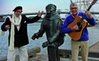 Foto på två män vid en staty som står alldeles vid vattnet i en hamn. Den ena spelar gitarr.