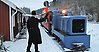 Bild på ett gammaldags tåg med ett blått lok. Det ligger snö på marken och en man vinkar in tåget till stationen.