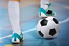 Foto på en persons ben med fotbollsskor. Ena foten står på en fotboll. Personen står på ett golv med olika linjer på i en sporthall.