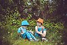 Foto på två barn med hattar som sitter ner i gräset och läser i en bok.