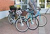 Tre cyklar uppställda utanför biblioteket i Munkedal. Två elcyklar med cykelkorg. På styret hänger en hjälm