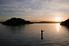 Solnedgång. Mitt ute i vattnet står en person och fiskar med ett flugfiskespö. Längre ut syns skogbeväxta öar.