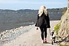 Foto på en kvinna med ljusblont hår klädd i svart som går på en grusväg mellan ett berg och havet tillsammans med en kopplad hund.
