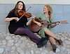 Två kvinnor sitter på en kullerstensgata lutade mot en vägg och spelar på gitarr och fiol.