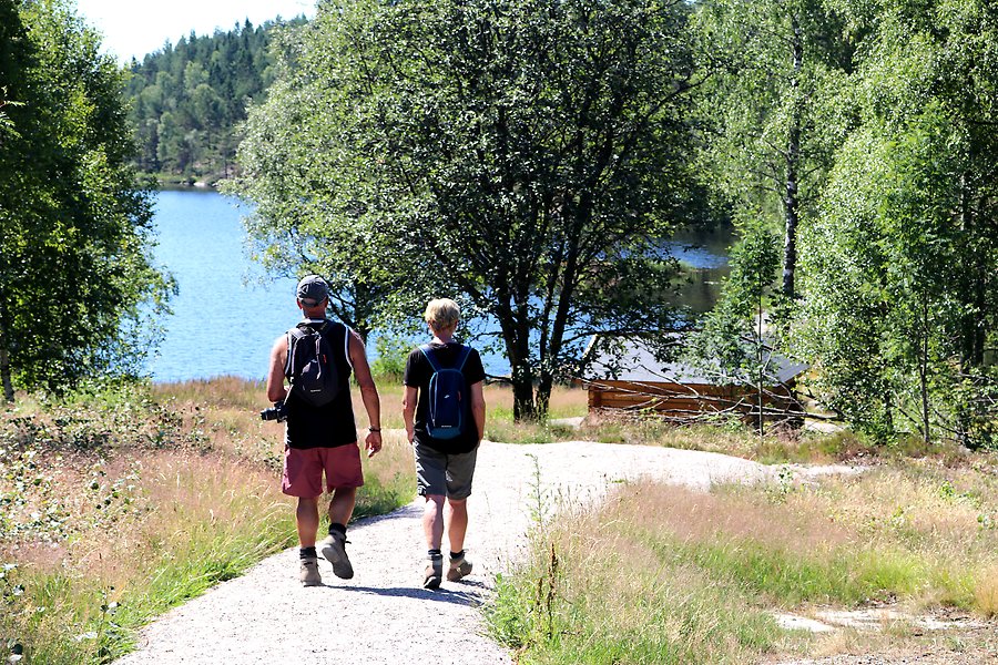Bild på två personer som går längs en promenadväg ner mot en sjö i ett lummigt skogsområde.
