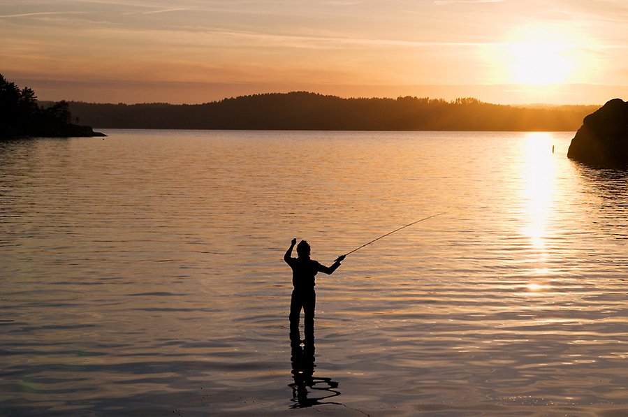 Foto på en fiskare som står i vattnet och fiskar med spö. Solen går ner och på vattnet finns krusningar. I bakgrunden ser man berg med växtlighet.