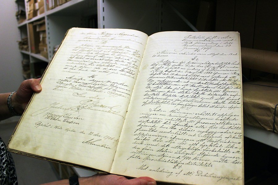 En person står i ett arkiv och håller upp en stor bok som innehåller protokoll från 1906.