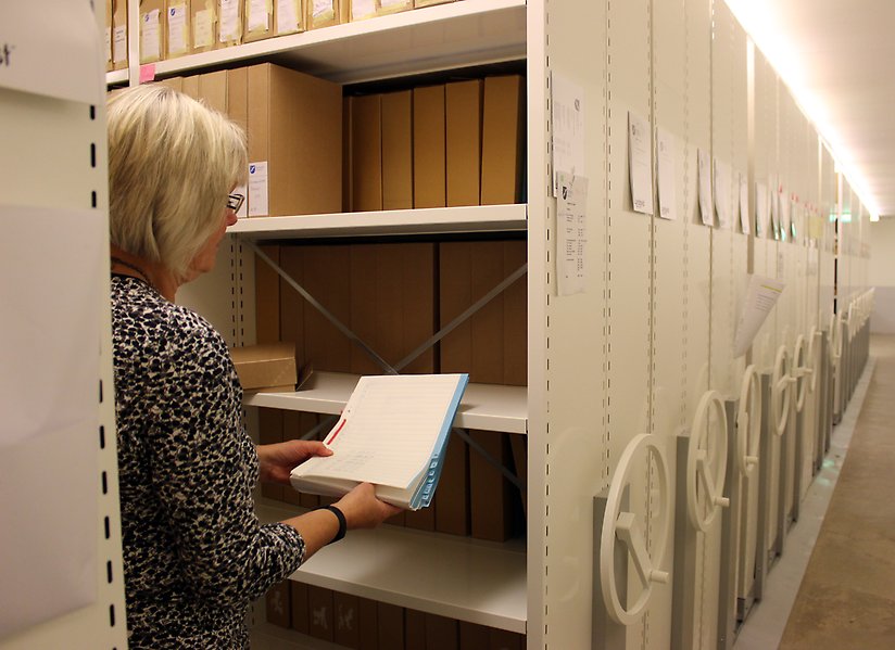 På bilden syns en person som håller upp en bunt dokument i ett rum med massor av hyllor fyllda av arkivlådor.