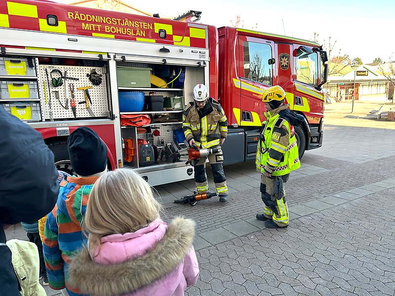 På ett torg står en brandbil. Vid brandbilen står barn och två brandmän. Brandmännen visar hur olika verktyg och detaljer på brandbilen fungerar.