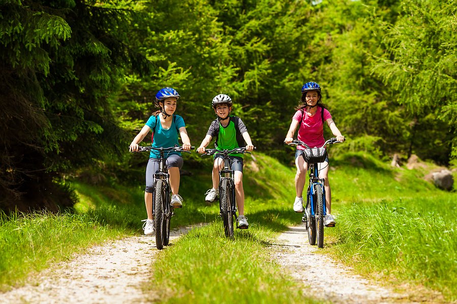 Tre personer som cyklar på en lite skogsväg. En vuxen kvinna tillsammans med en flicka och en pojke. Alla personer bär cykelhjälmar.