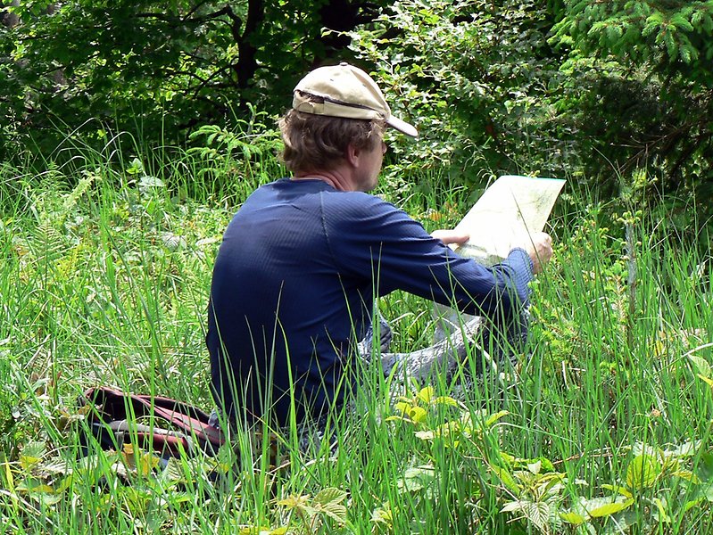 En man sitter i högt grönt gräs och studerar en karta. Solen lyser.