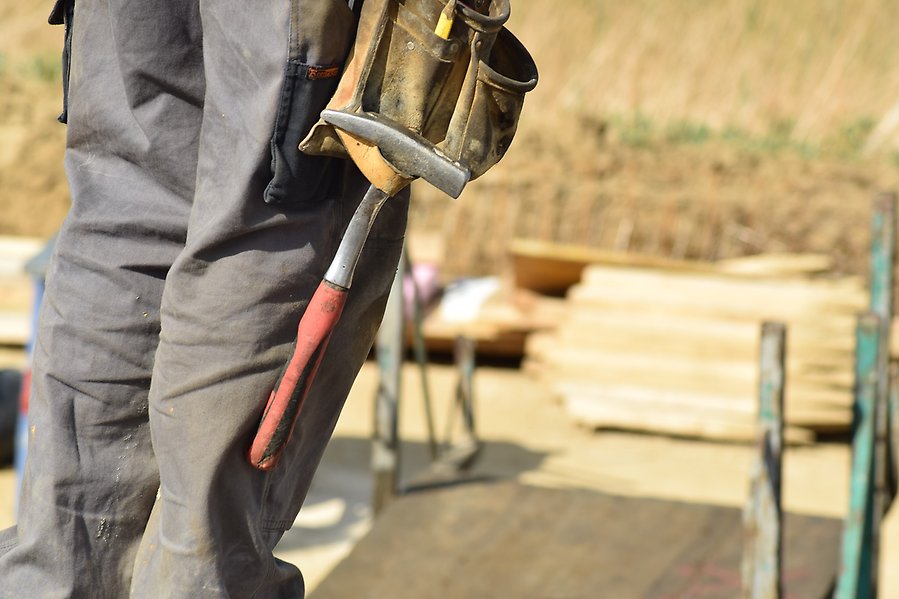 Foto på en persons ben i arbetsbyxor med hammare i verktygsbälte. I bakgrunden syns staplade skivor i trä.