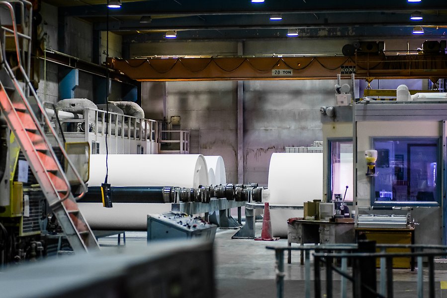 Foto i en industrilokal med maskiner och stora vita pappersrullar.