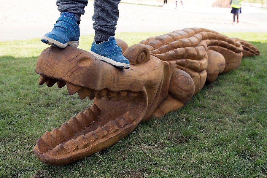 Bild på en krokodil skulpterad i trä. På krokodilens huvud står ett barn, endast fötterna syns i bild.