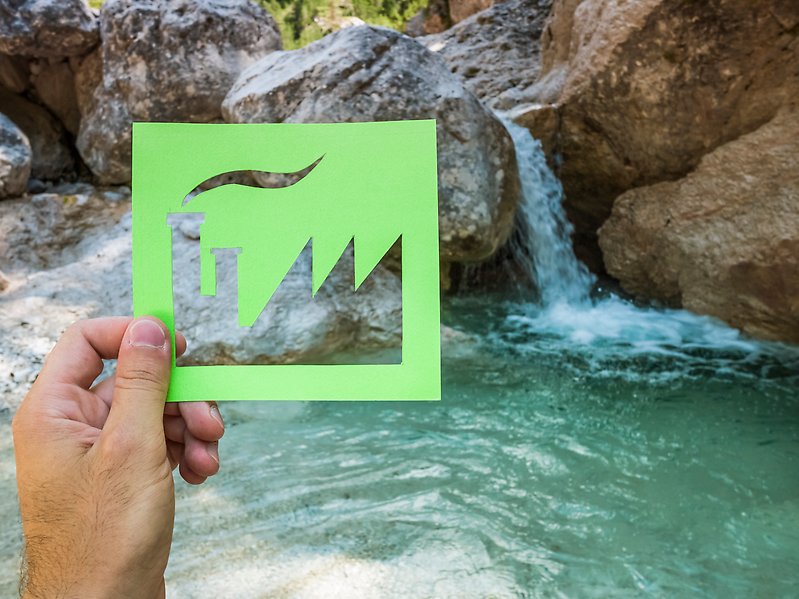 Foto på en hand som håller i en illustration av en fabrik i grönt framför ett vattenfall.