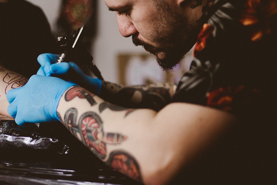 Foto på en man som tatuerar en person. Mannen har tatueringar på armen och plasthandskar på händerna.