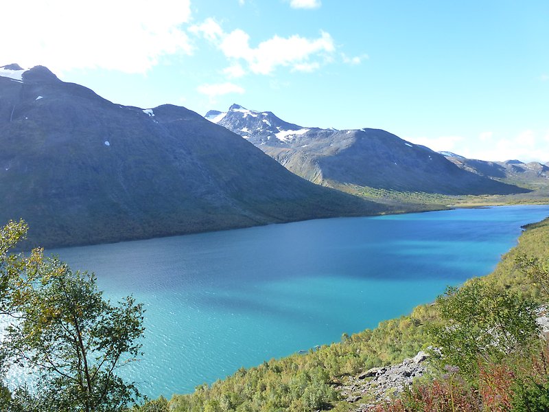 Foto på en turkos sjö med höga blånande berg i bakgrunden. I förgrunden syns en grön sluttning.