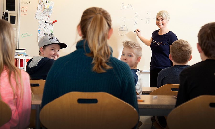 En lärare står framme vid tavlan i ett klassrum. Två av barnen tittar nyfiket bakåt mot fotografen.