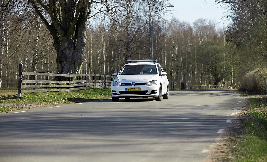 På en landsväg omgiven av lövträd och delvis trädgårdsstaket kör en vit personbil med Västtrafiks logga på.