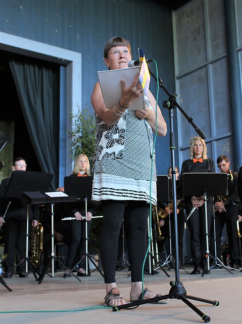 Foto på en kvinna som står vid en mikrofon på en scen och håller ett papper framför sig. I bakgrunden syns delar av en orkester.