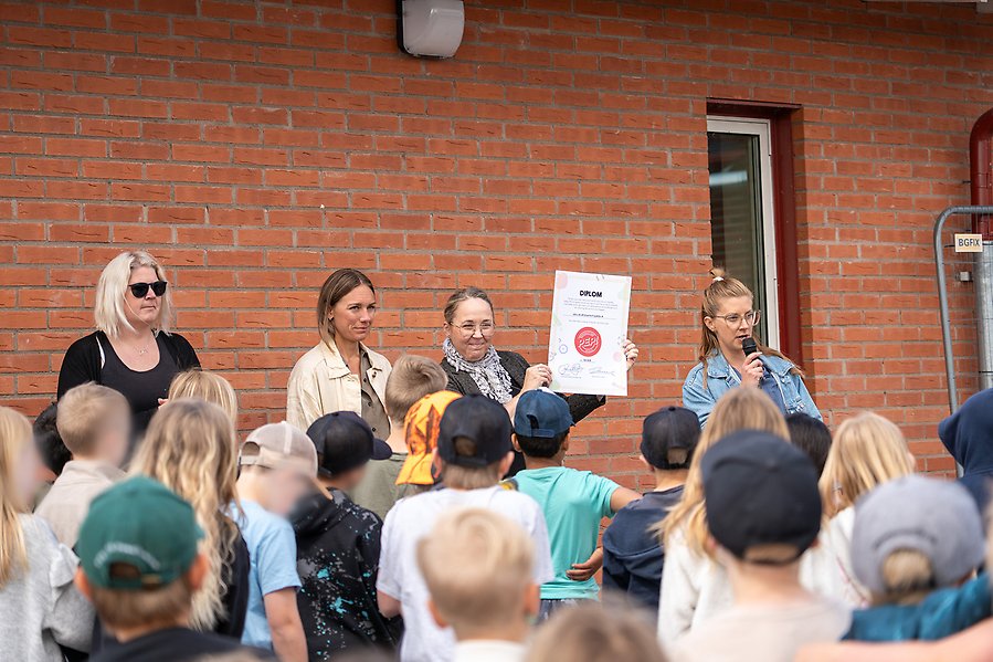 Fyra kvinnor står på en scen varav en håller upp ett diplom. Det står barn i publiken.