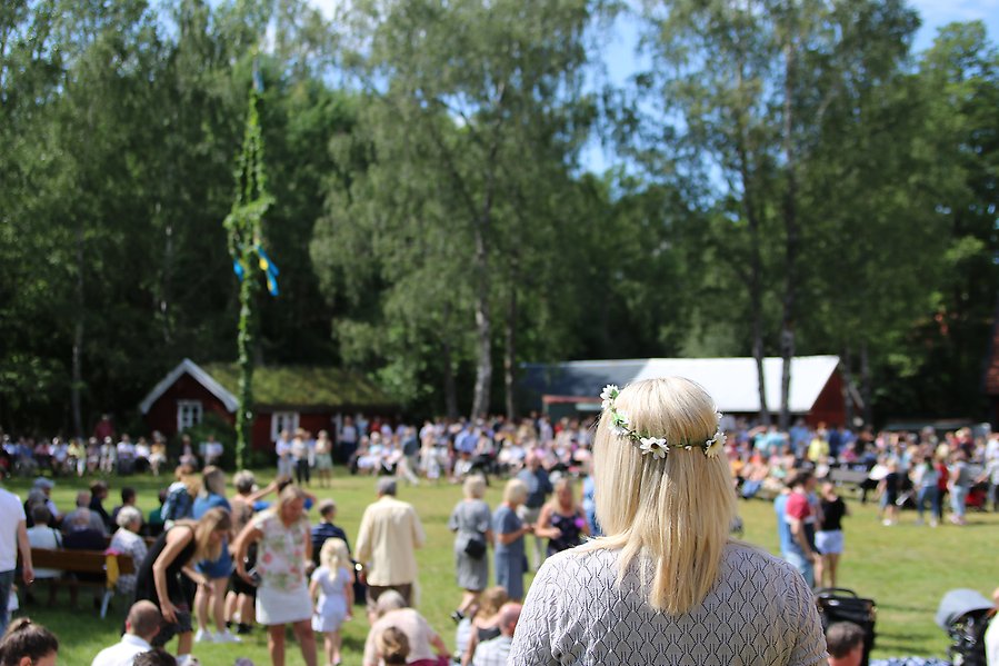 EN tjej som står och tittar på en midsommarstång. I bakgrunden syns det människor.
