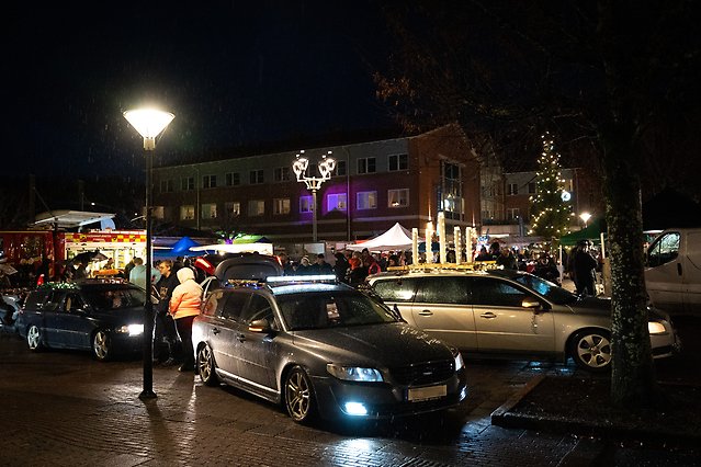 Bilar parkerade på ett torg. I bakgrunden syns en julgran som lyser.
