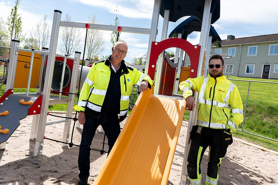 Två män i gula arbetsjackor står vid en klätterställning med en rutschekana. 