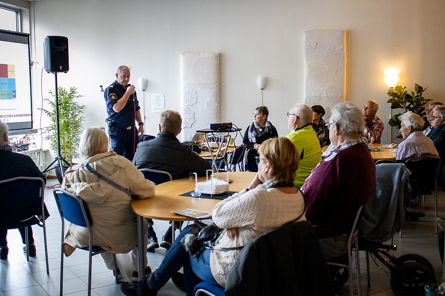 En polis som står med en mikrofon inför en samling personer som sitter ner.