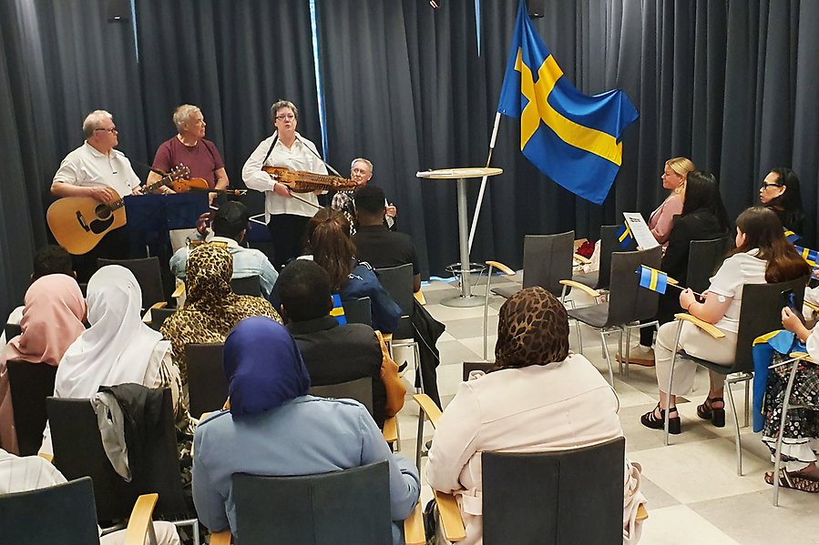 Medborgarskapsceremoni med sittande publik, tre som spelar instrument och sjunger samt en svenska flaggan i bild.